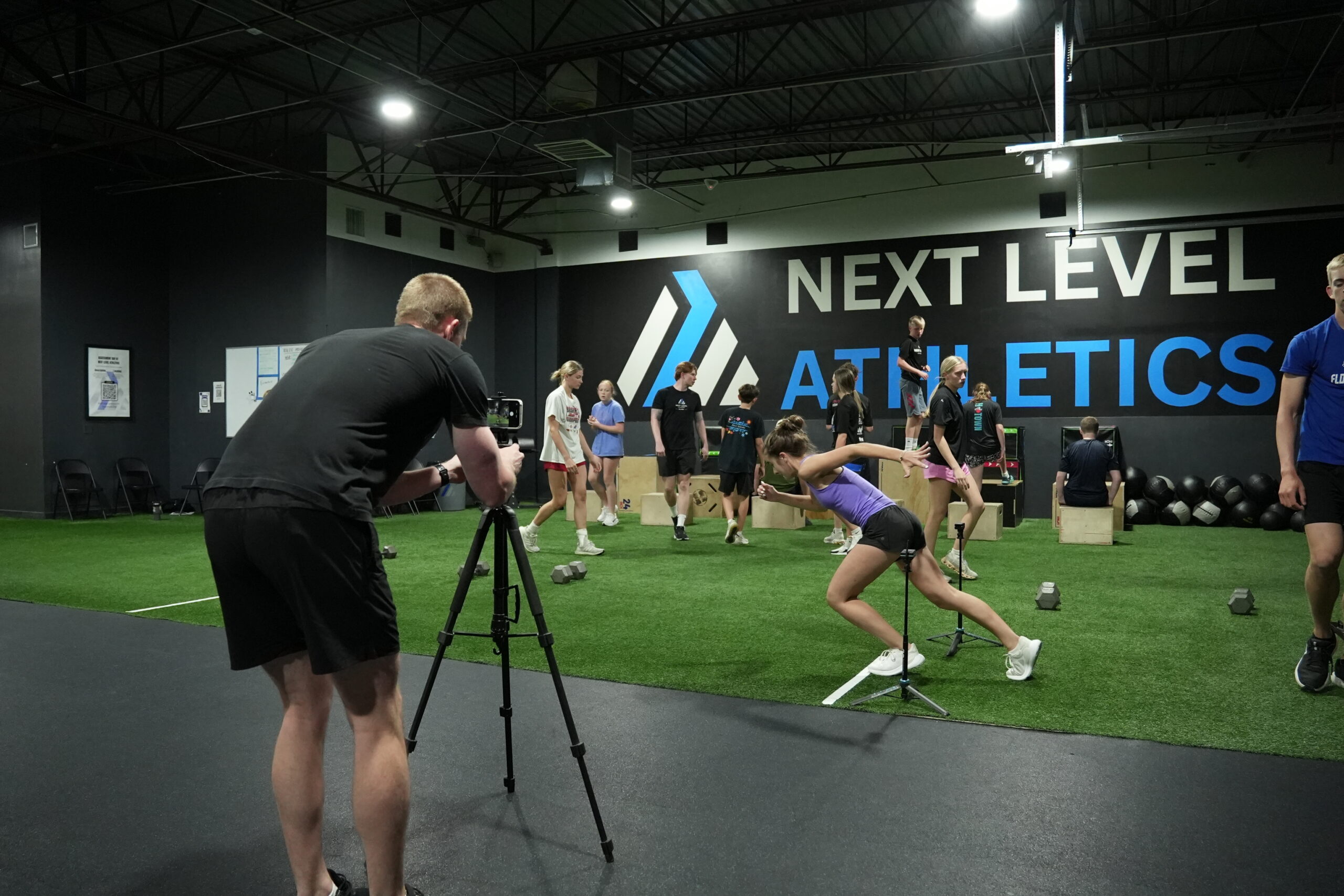 Sweets films an NLA athlete in sprint-start position during the Baseline Assessment, with the Next Level Athletics wall in the background